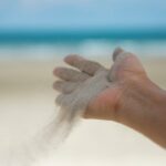 Close-up of hand releasing sand on sandy beach with ocean in background.