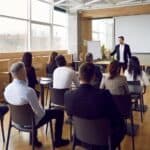 Team of people having class with business trainer. Group of male and female employees sitting at desks in modern office and listening to lecture by experienced teacher sharing knowledge and guidance