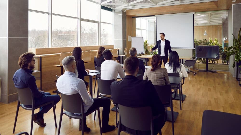 Team of people having class with business trainer. Group of male and female employees sitting at desks in modern office and listening to lecture by experienced teacher sharing knowledge and guidance