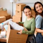 Young happy women having fun while carrying cardboard boxes into their new home.