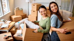 Young happy women having fun while carrying cardboard boxes into their new home.