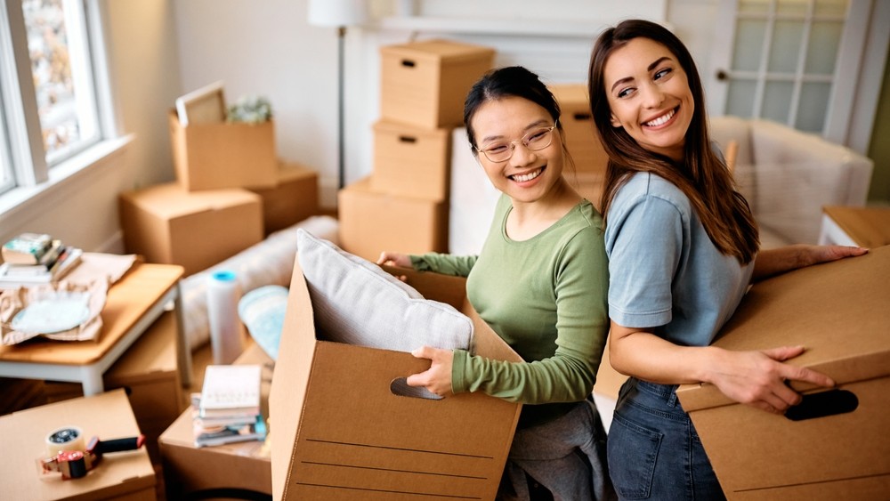 Young happy women having fun while carrying cardboard boxes into their new home.