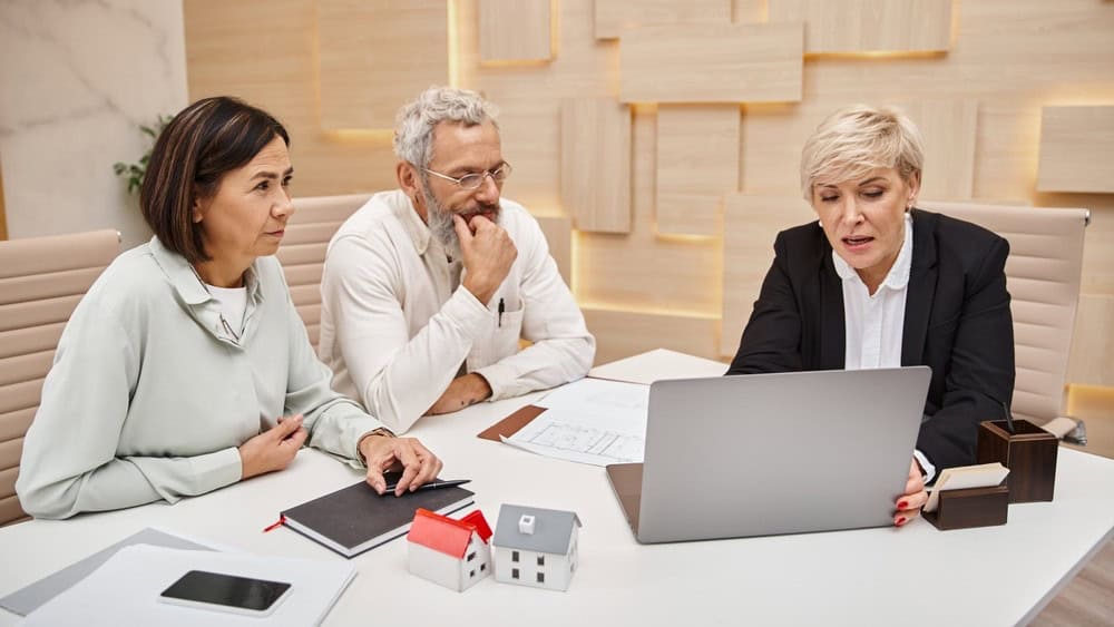 happy middle aged married couple looking at female realtor showing project of new home on laptop
