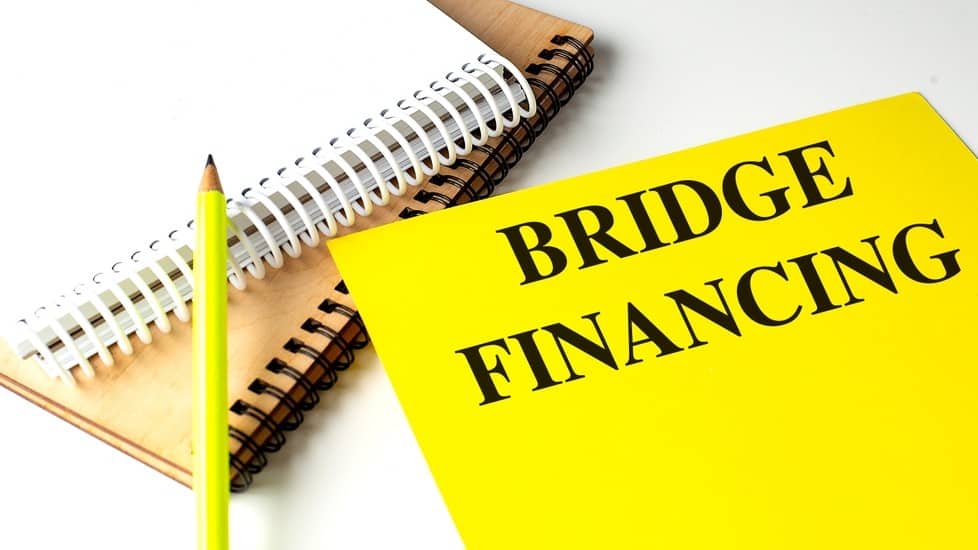 Yellow folder with words bridge financing next to notebooks and pencil on white background .