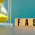 Yellow hourglass beside wooden blocks spelling FAST, representing speed, efficiency, productivity, quick decisions, time management, business performance, and rapid execution.